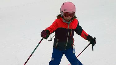 Child in blue ski pants, black and red jacket and pink helmet skis down a snowy hill.
