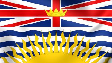 The B.C. provincial flag showing the union jack at the top, at the centre of which is the crown of King Edward and the bottom of the flag shows wavy blue lines representing the Pacific ocean with a yellow setting sun at the bottom.